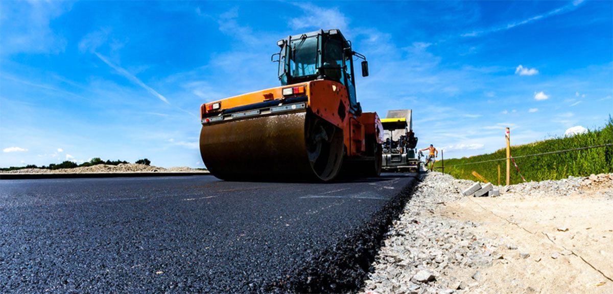 Road roller paving fresh black asphalt beside a gravel shoulder under a blue sky
