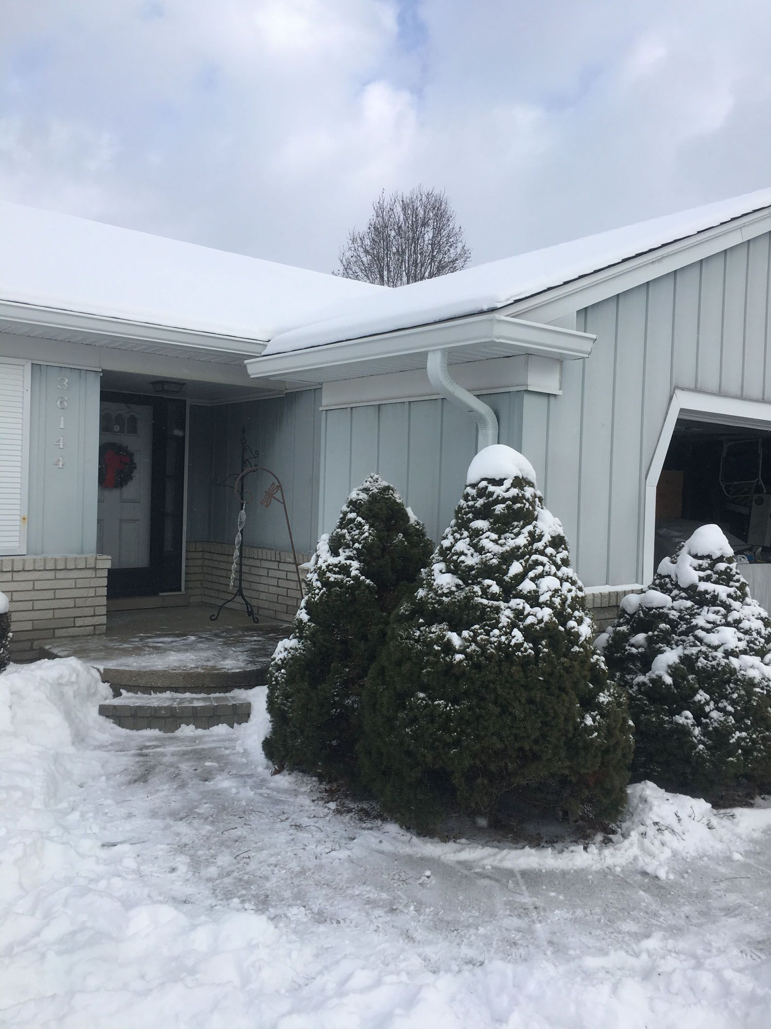 Snow-covered house entrance. Evergreen shrubs, steps, icy walkway. Cloudy sky.