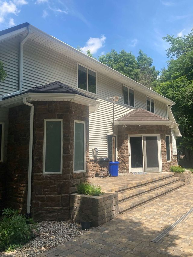 Two-story house with stone facade, beige siding, and multiple windows. Paved patio area with steps and landscaping.