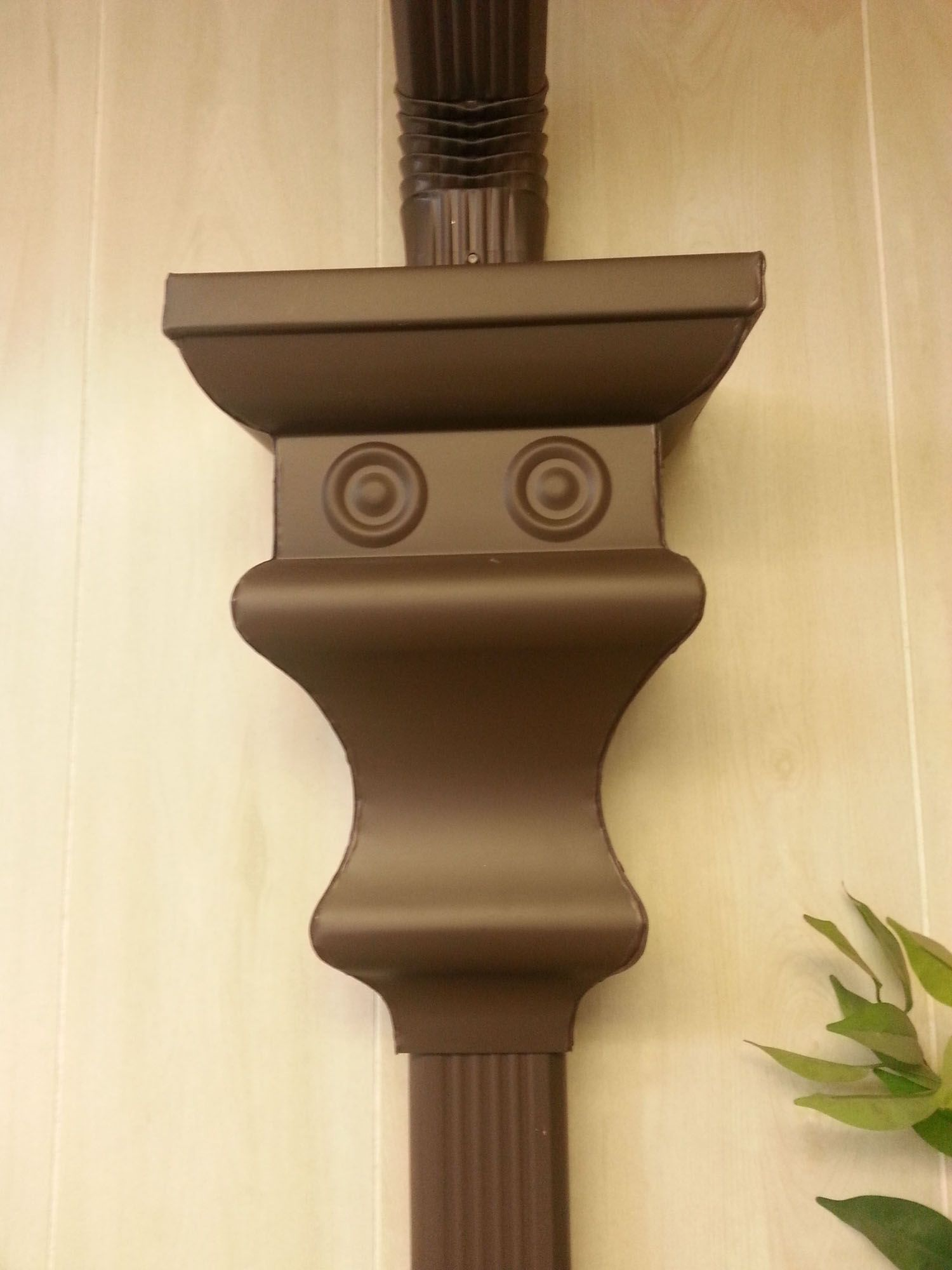 Brown decorative rain gutter downspout attachment against a beige wall, with two circular patterns.
