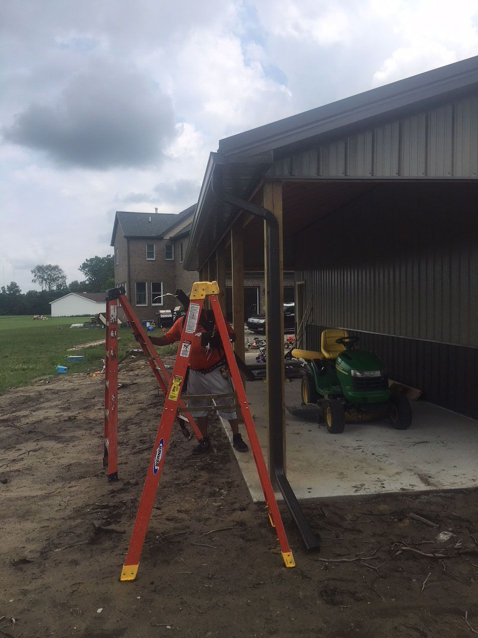 Person on ladder installing gutter on a building with a lawn tractor parked inside.