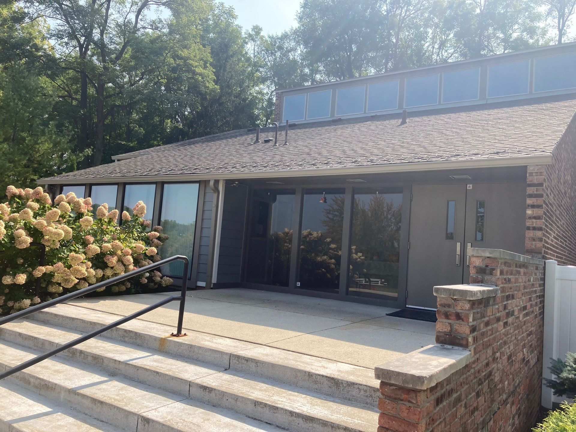 Building with windows and brick accents, steps lead to the entrance. Lush bushes and trees in background.