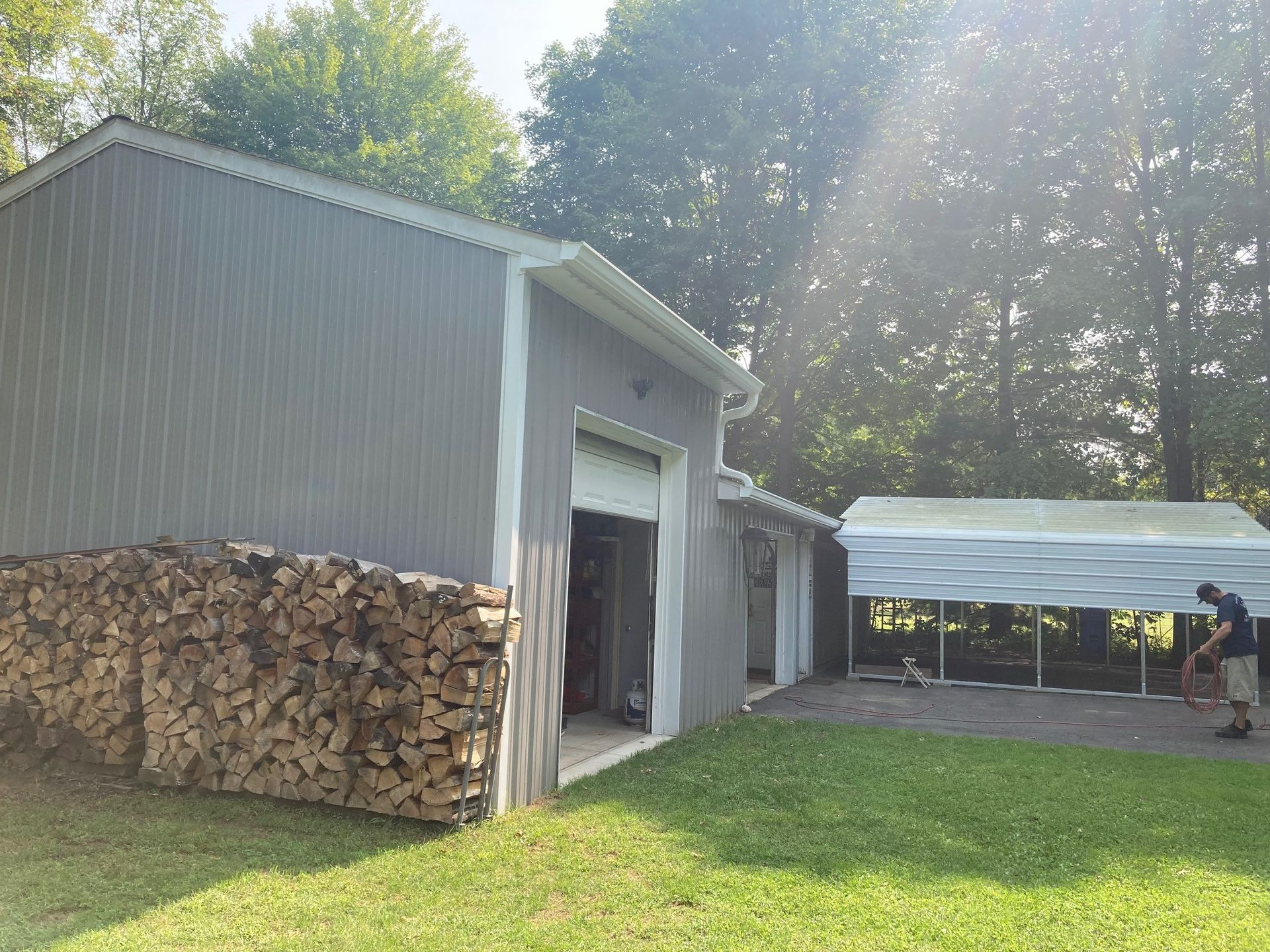 Gray garage with open door, stacked firewood, and a person near a white structure on grass. Sunny outdoors.
