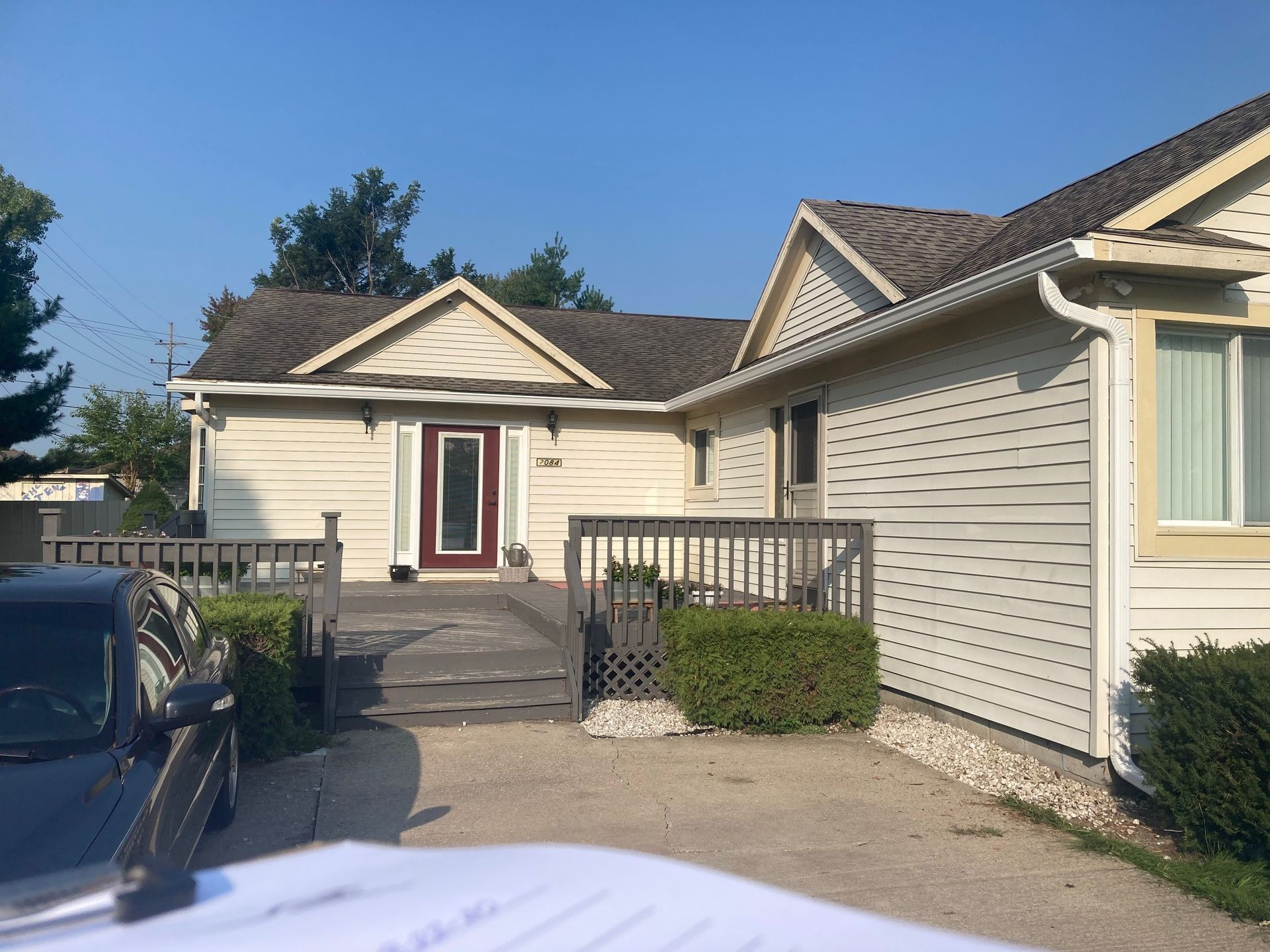 Beige house with a wooden deck and red door. Gravel driveway, blue sky.