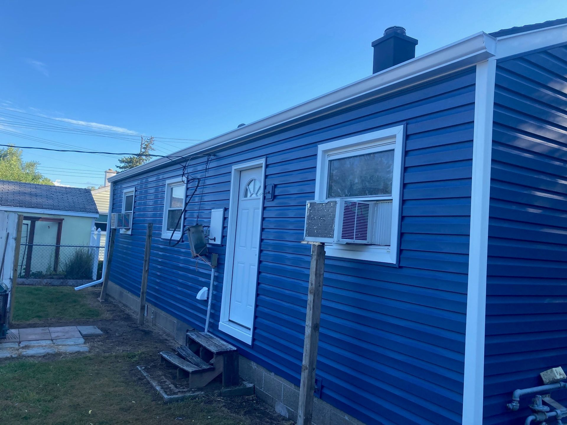 Blue-sided house with white trim, windows, and door. An air conditioner is in the window, with grass and a fence in the yard.