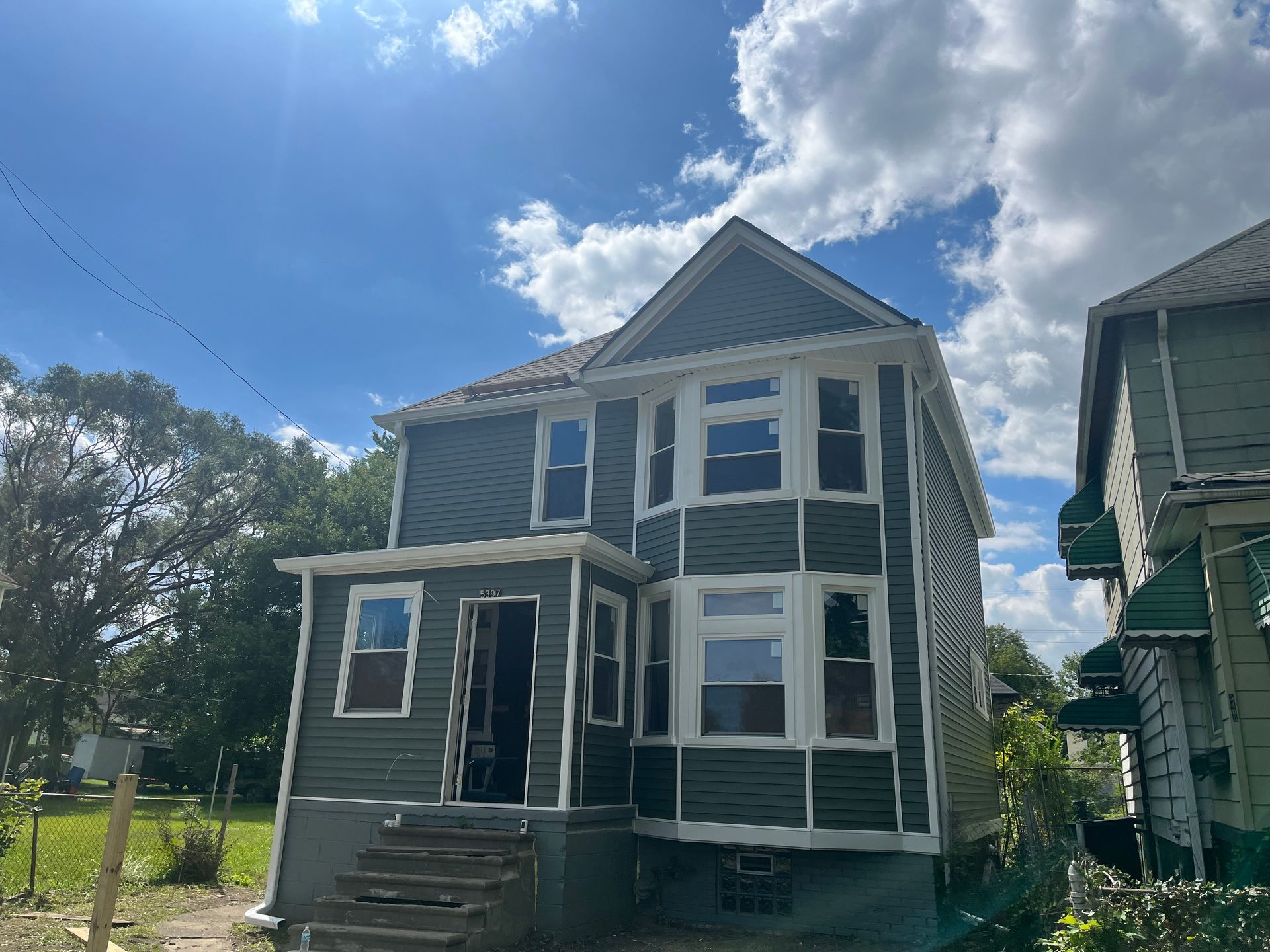 Green two-story house with bay windows and a porch under a partly cloudy sky.