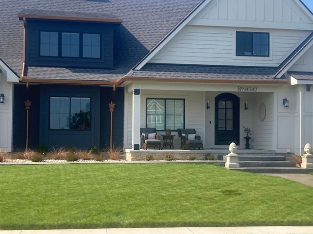 Modern two-story house with white siding and dark blue accents. Green lawn, porch with seating, and dark front door.