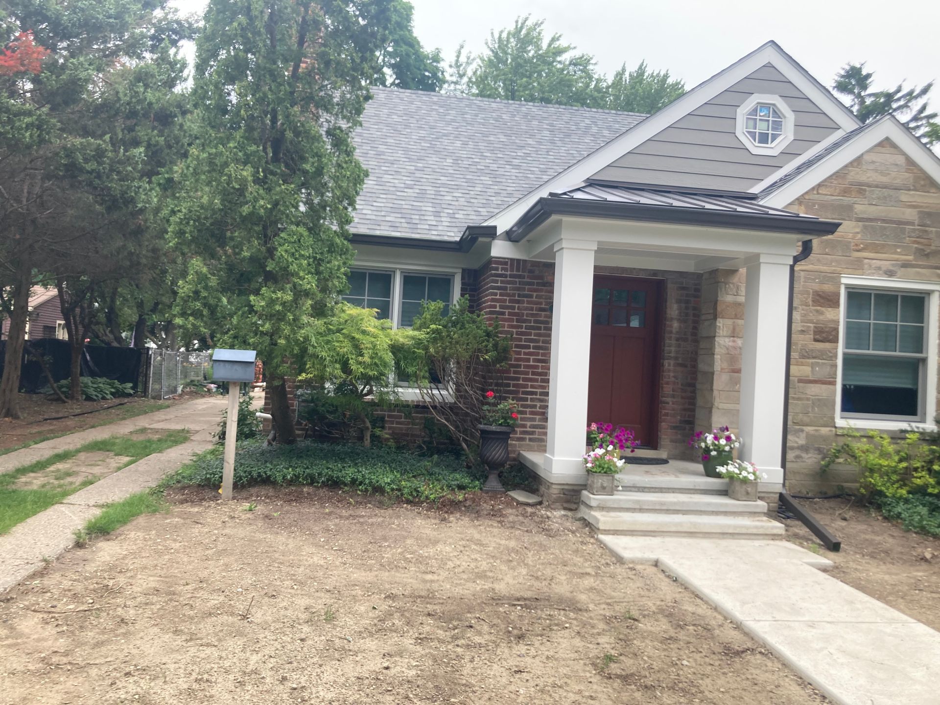 House exterior: stone and brick facade, red door, gray roof, concrete walkway, mailbox, and trees.