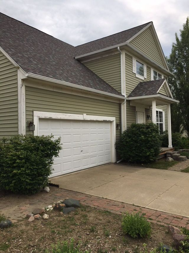 Two-story house with green siding, white trim, and a brown roof. White garage door, bushes, and a driveway.