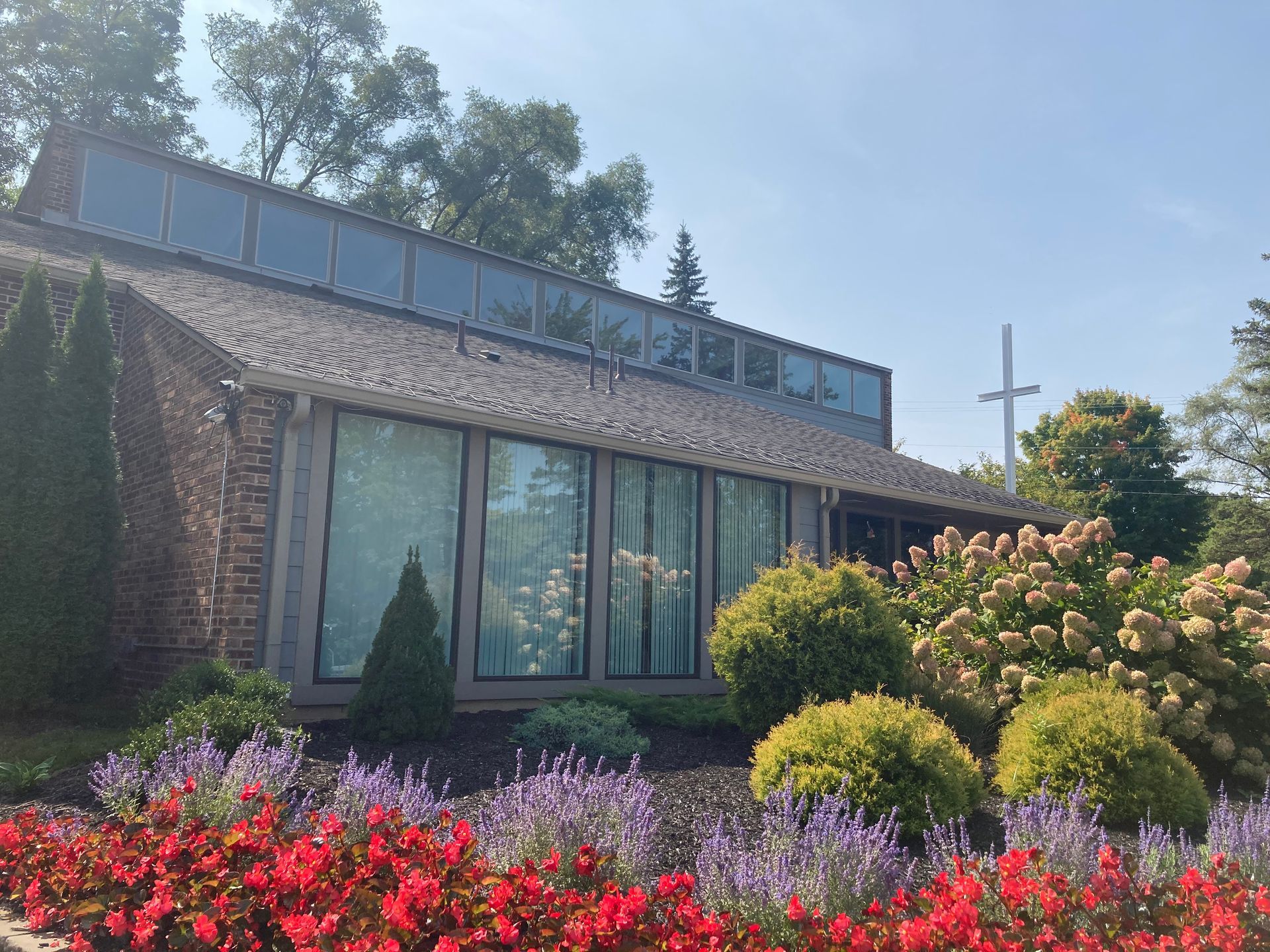 Exterior of church with large windows, cross, and colorful flower beds.