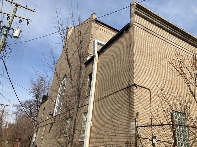 Tan brick building with a tall, arched window and bare trees against a blue sky.