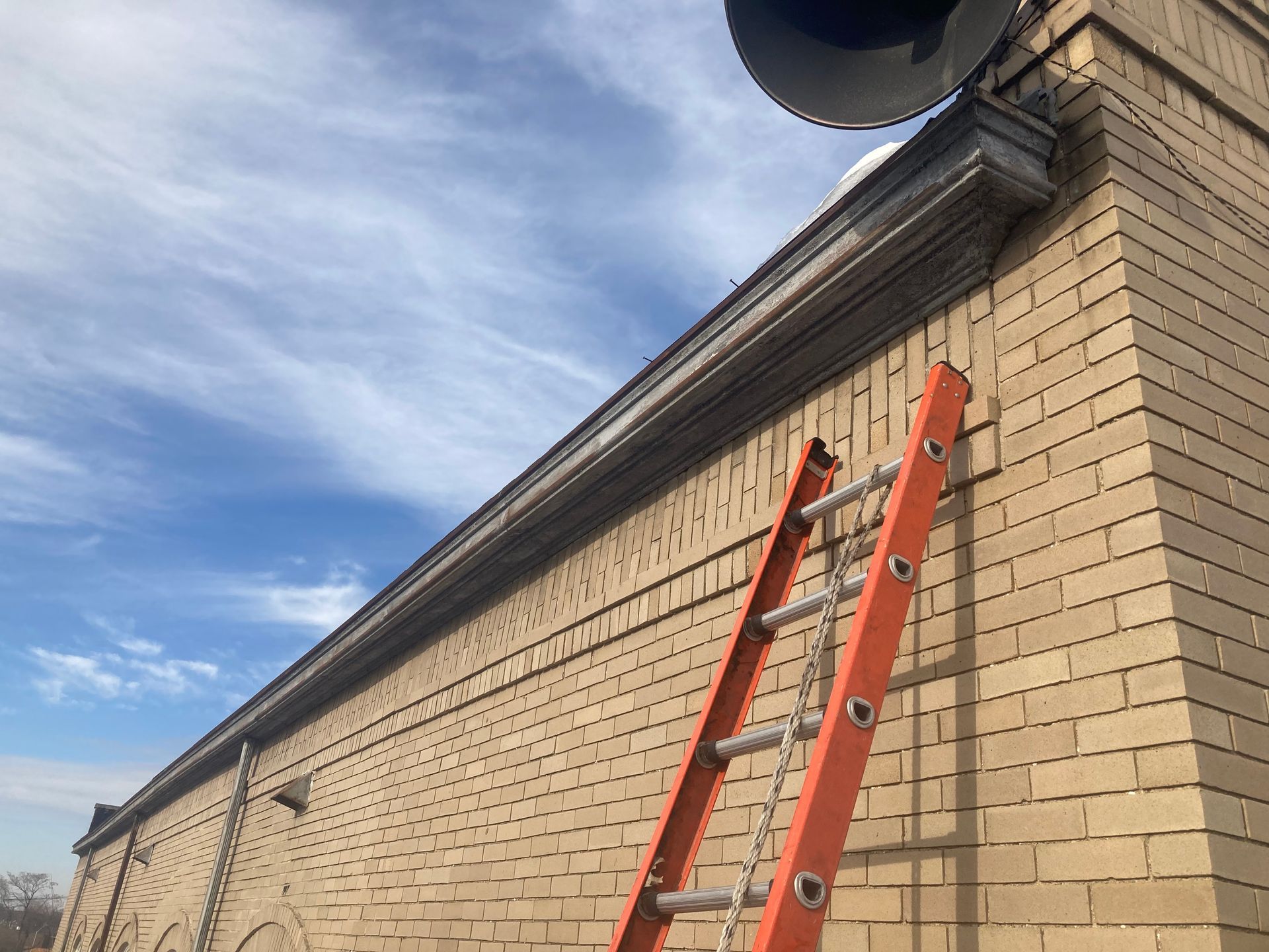 Orange ladder leaning against a light brick building. Blue sky with clouds.