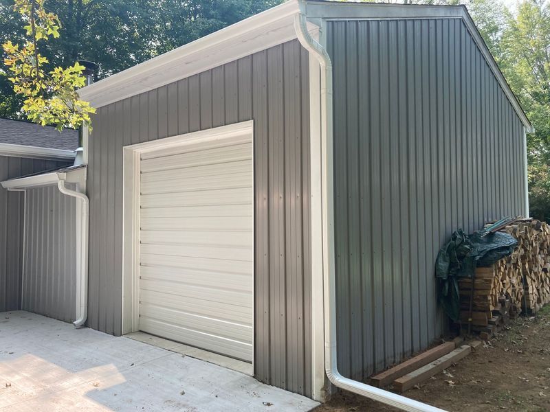 Gray and white garage with a white garage door, surrounded by stacked firewood.