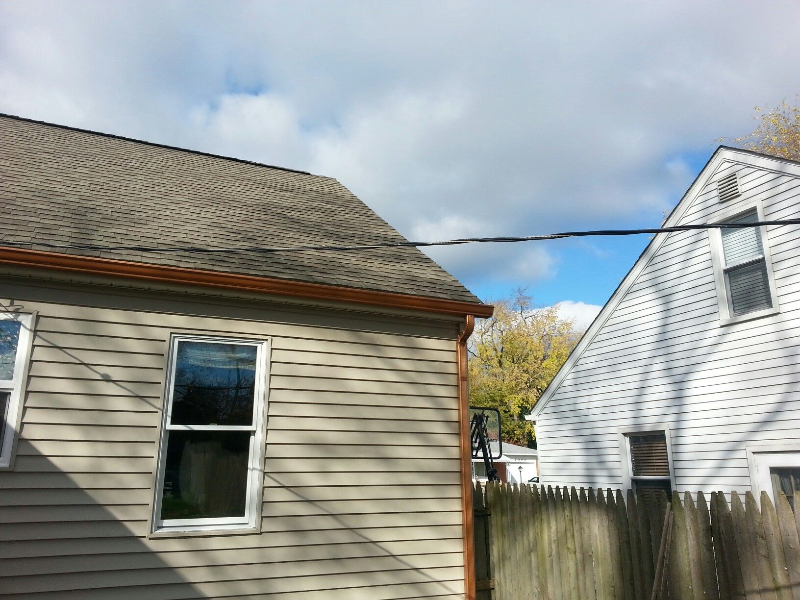 Two houses with a utility line, brown gutters, and a wooden fence under a cloudy sky.