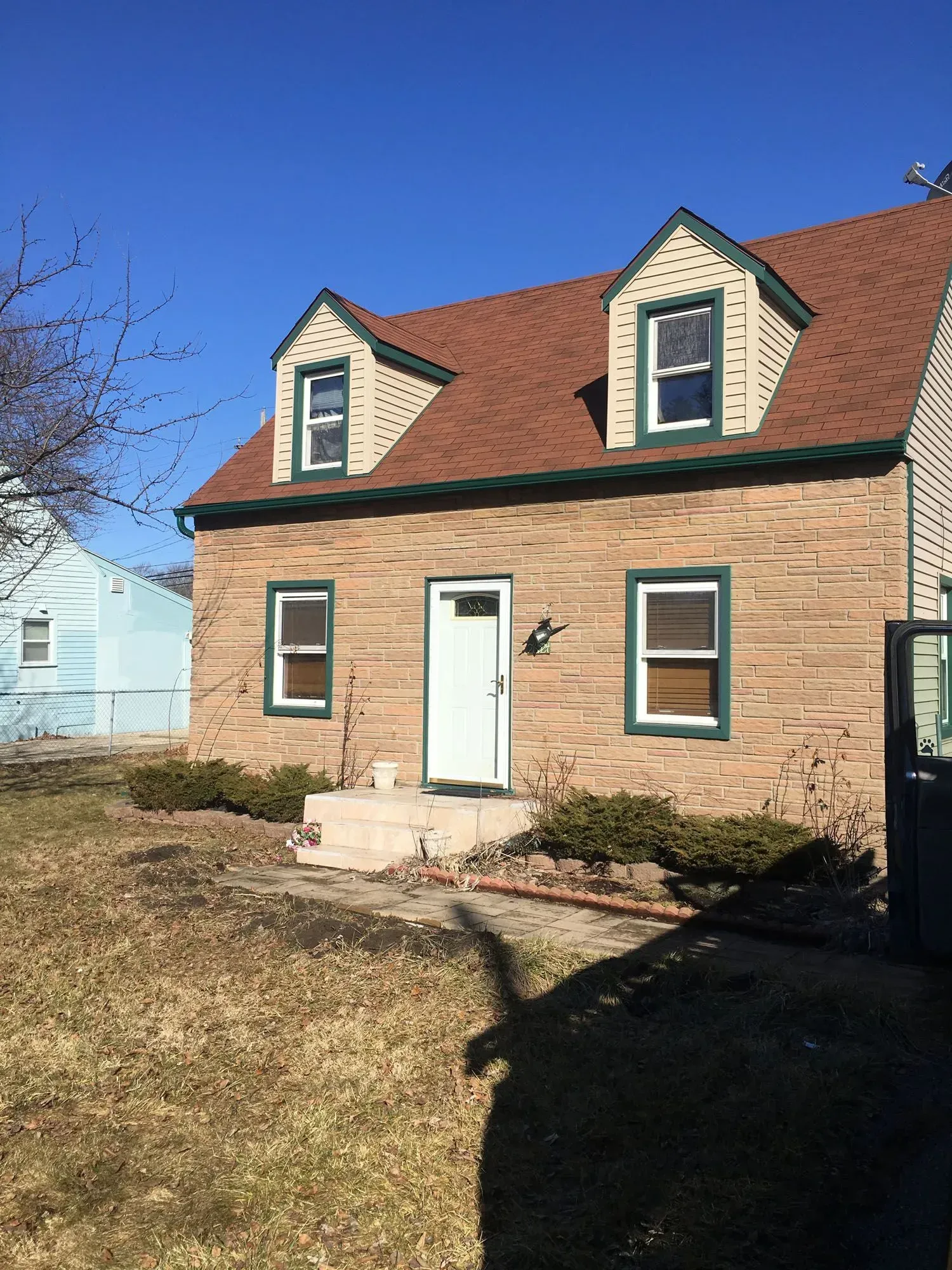 Brick house with red roof, two dormers, green trim, white door, and small front yard with dry grass.