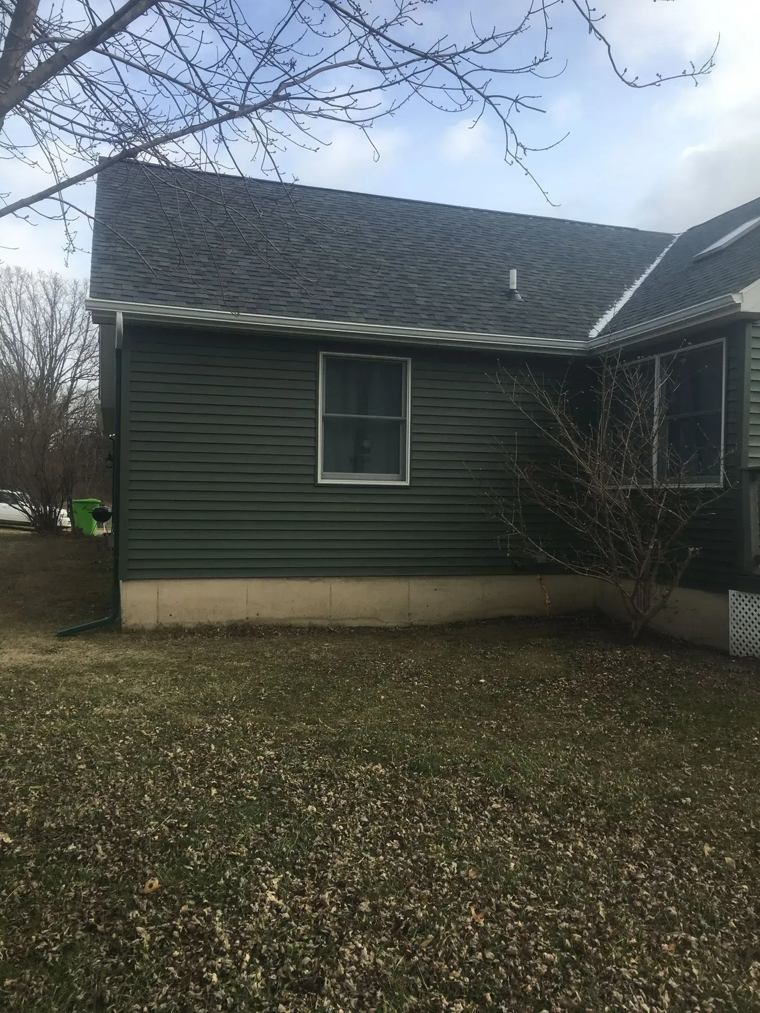 Green-sided house with a dark roof and a window. Overcast day, dry grass.