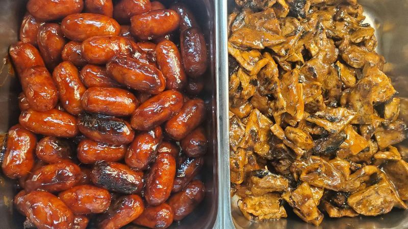 Two trays of food , one filled with sausages and the other with meat.