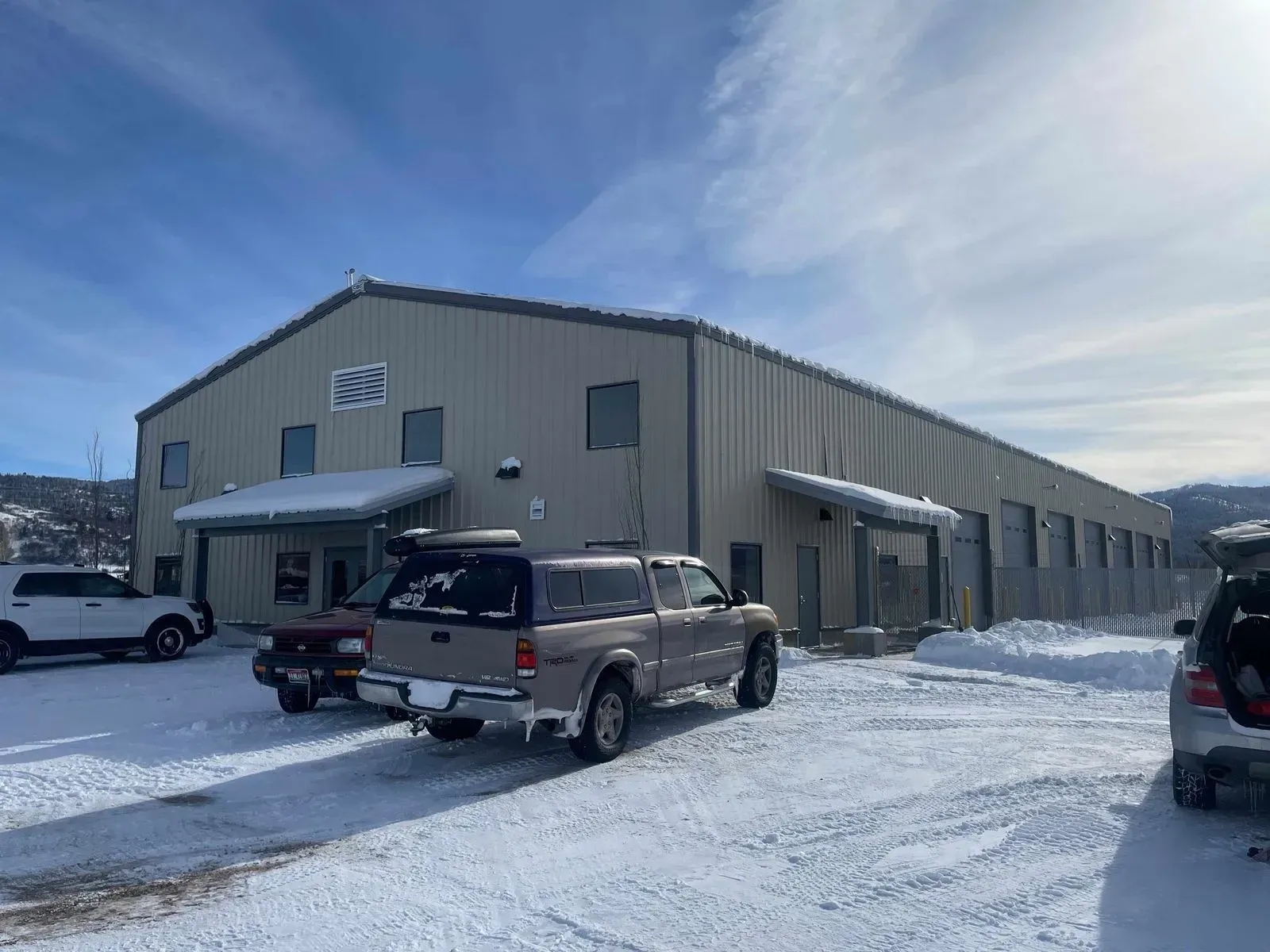 Exterior view of a large industrial building with snow and parked vehicles under a bright, partly cloudy sky