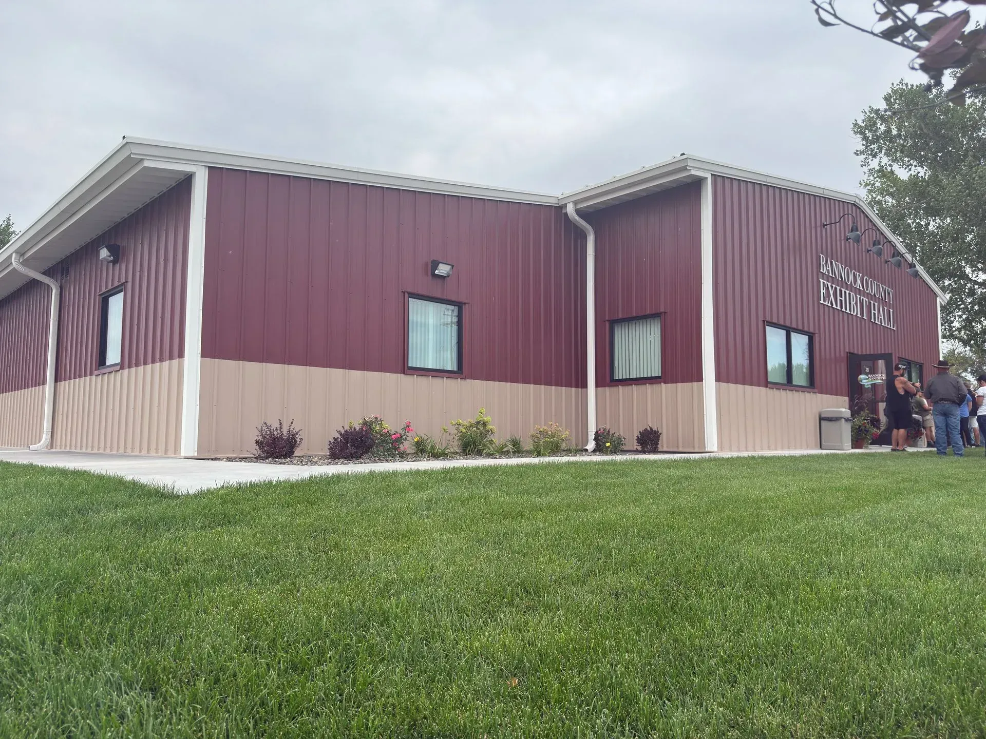 Red and tan metal building with small windows, a concrete walkway, and a grassy lawn.