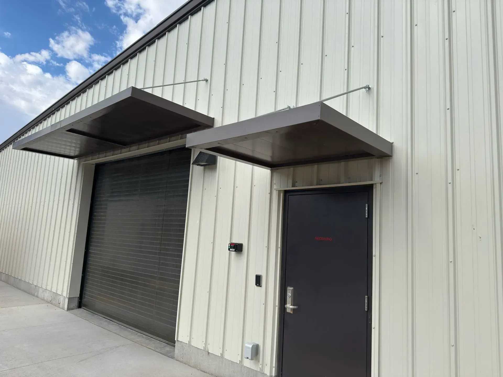 Warehouse exterior with a roll-up door and entry door, both with brown awnings. Light siding, blue sky.