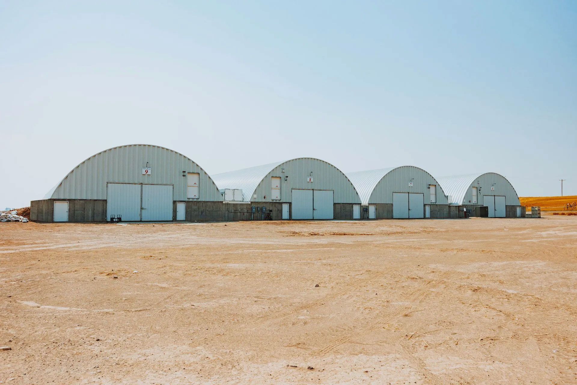 Four arched metal buildings in a desert landscape under a clear, blue sky.
