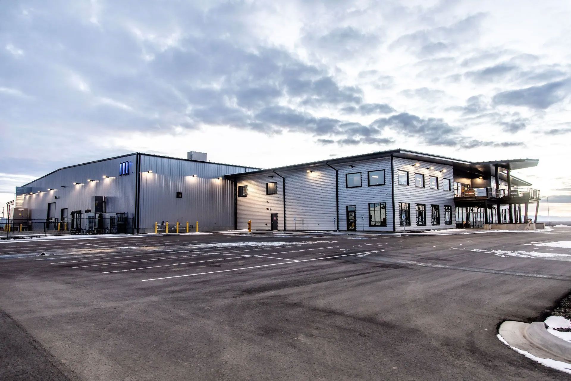 Large modern industrial building with a gray exterior and a large parking lot under a cloudy sky.