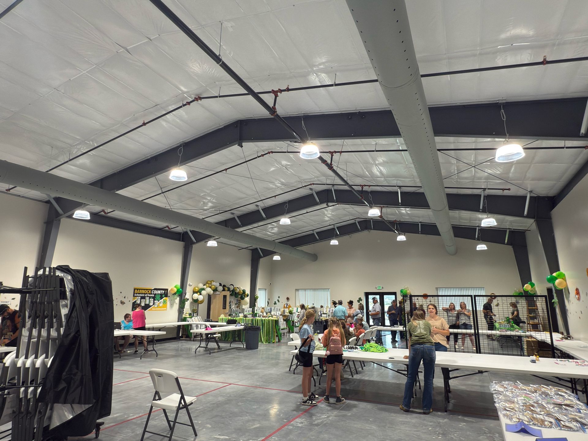 Large indoor event with tables, people, green decorations, and overhead lights.