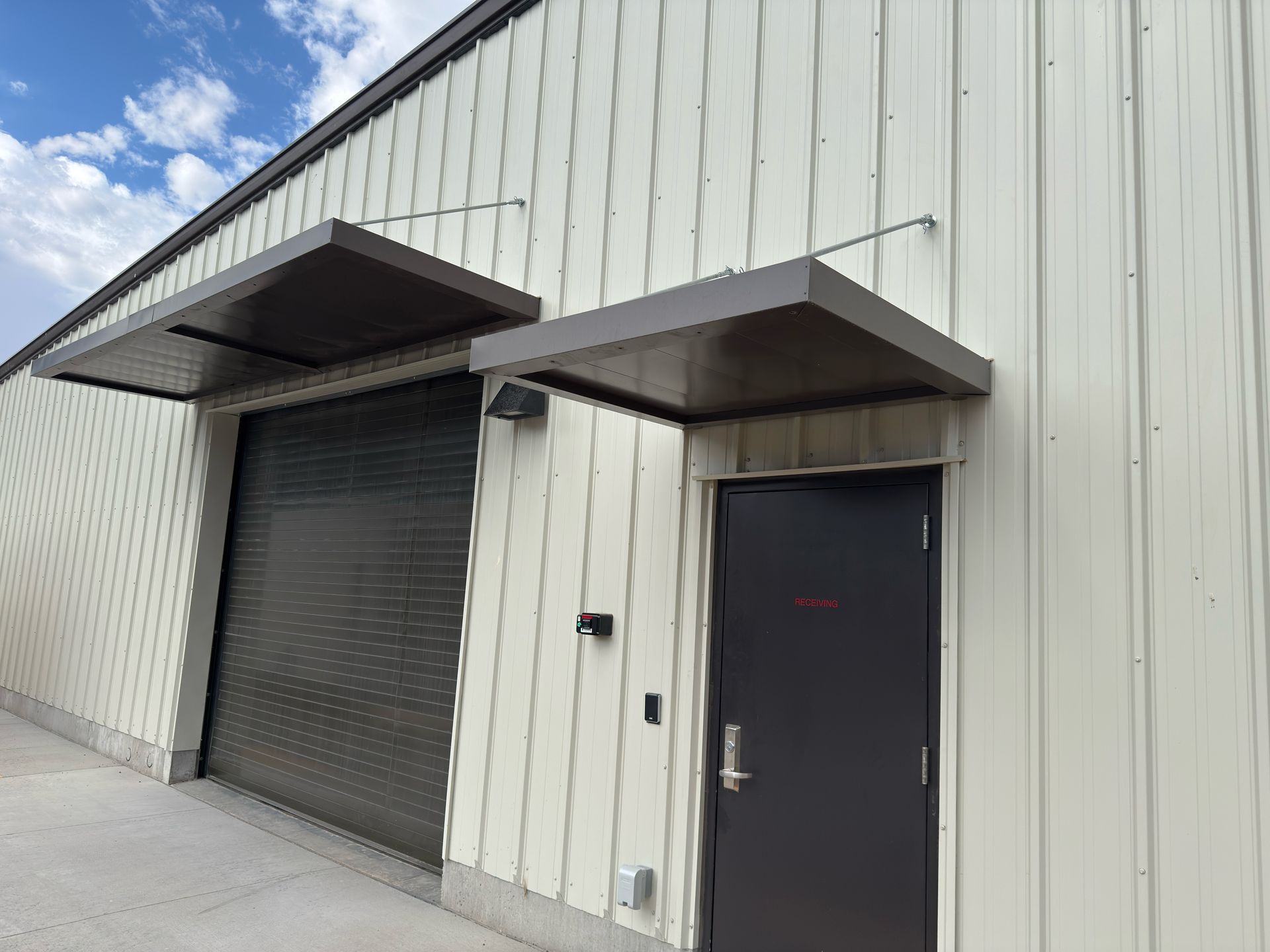 Warehouse exterior with a roll-up door and entry door, both with brown awnings. Light siding, blue sky.