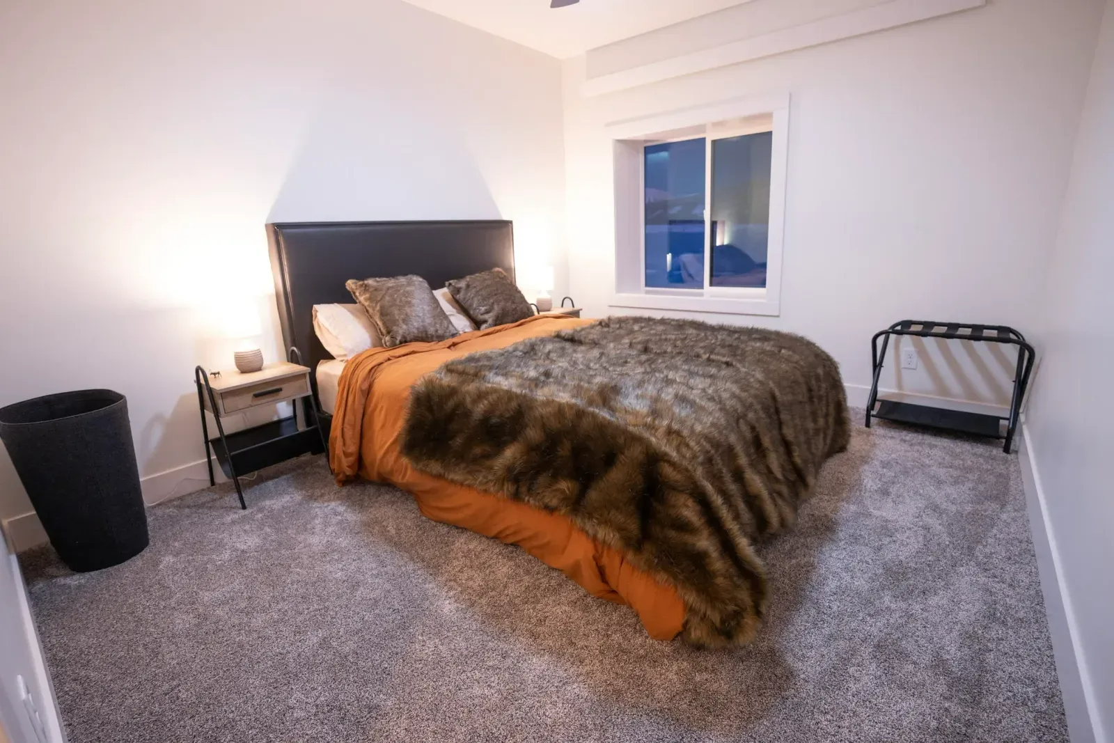 A bedroom with a bed , nightstand , trash can and window.