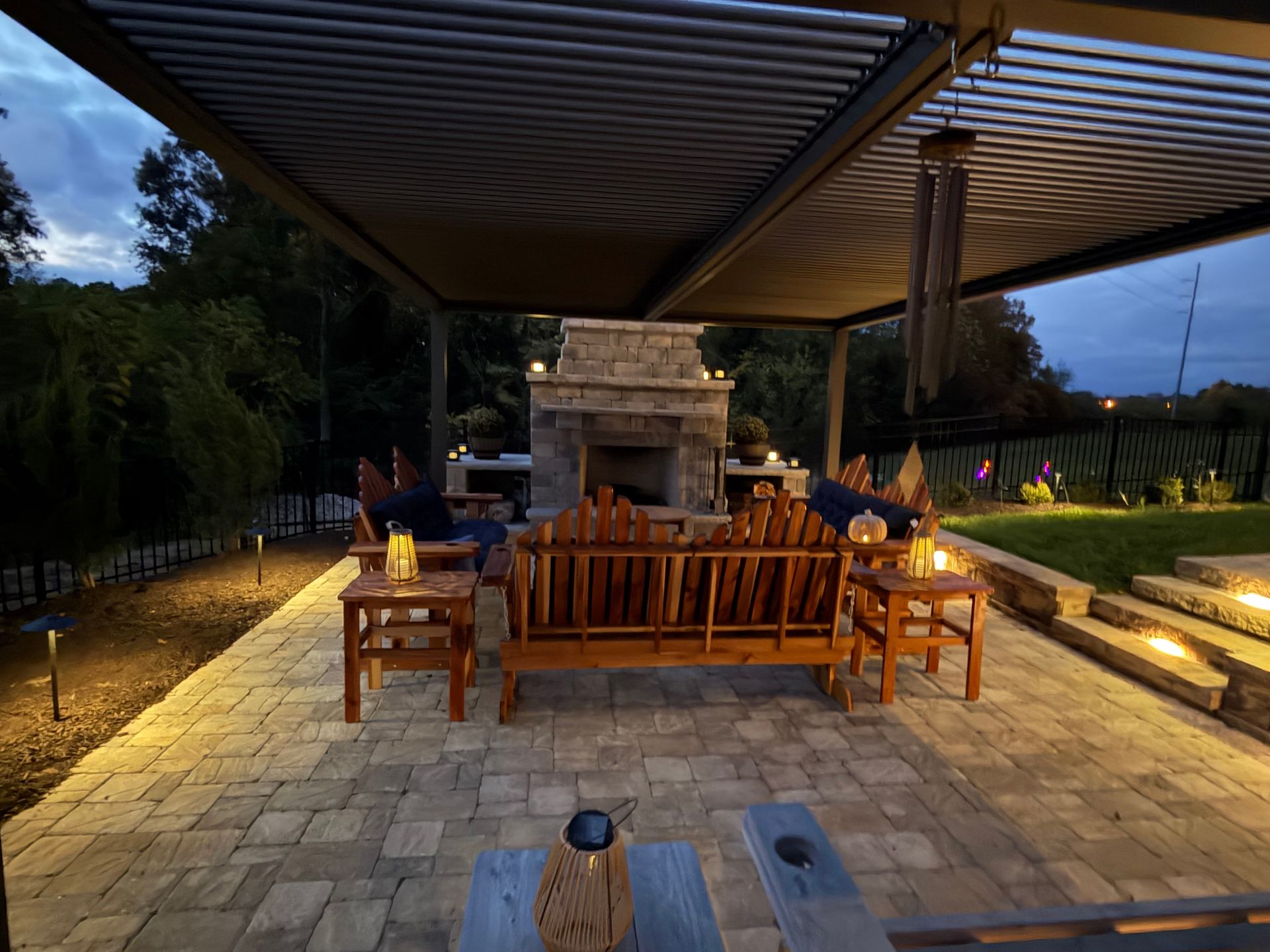 Patio with fireplace, seating, and overhead pergola at dusk; lit by warm lights.