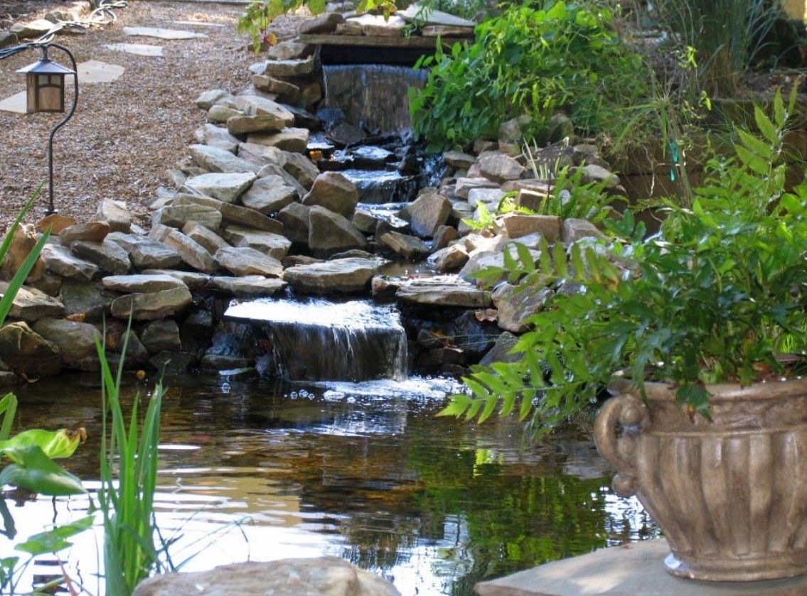 Waterfall cascading into a pond, surrounded by rocks and lush greenery. A potted fern sits nearby.