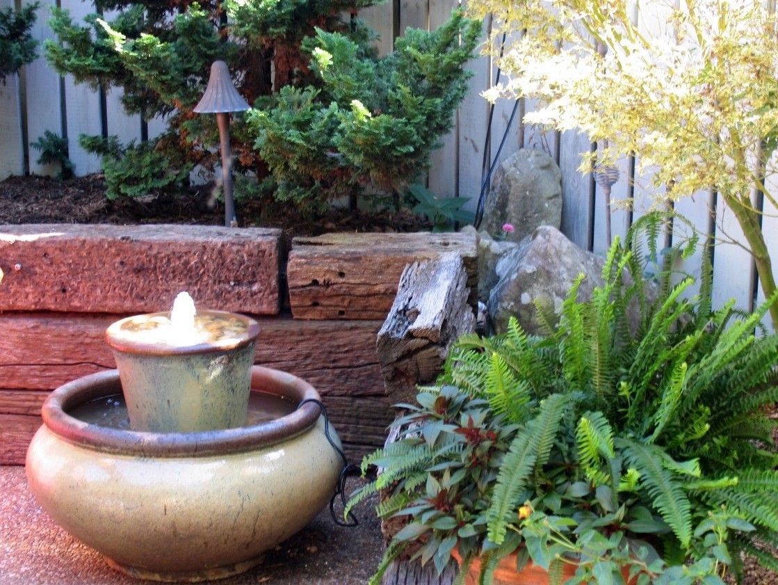 Water fountain in a ceramic pot, surrounded by greenery and a rustic wooden backdrop.
