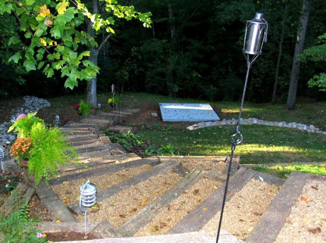 Stone steps lead down to a grassy yard with a metal torch and a white rectangular structure. Green foliage surrounds.