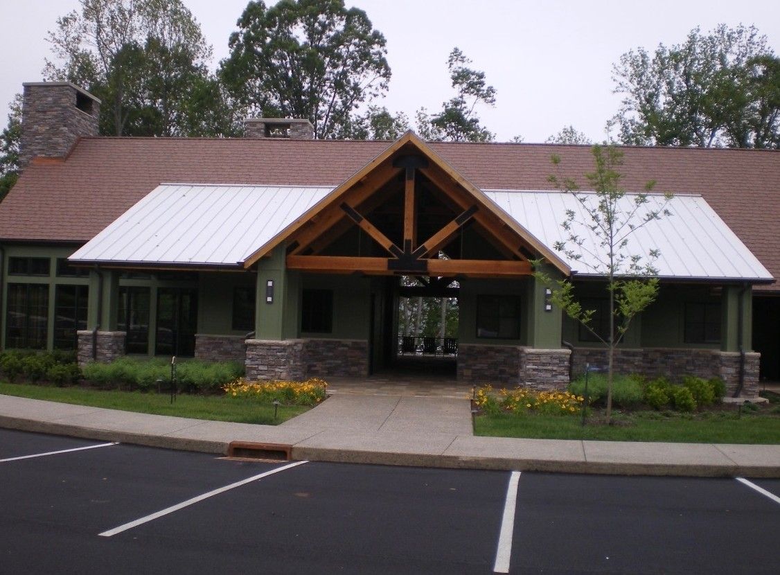 Green building with stone accents, brown roof, and metal awning. Paved walkway and parking area.