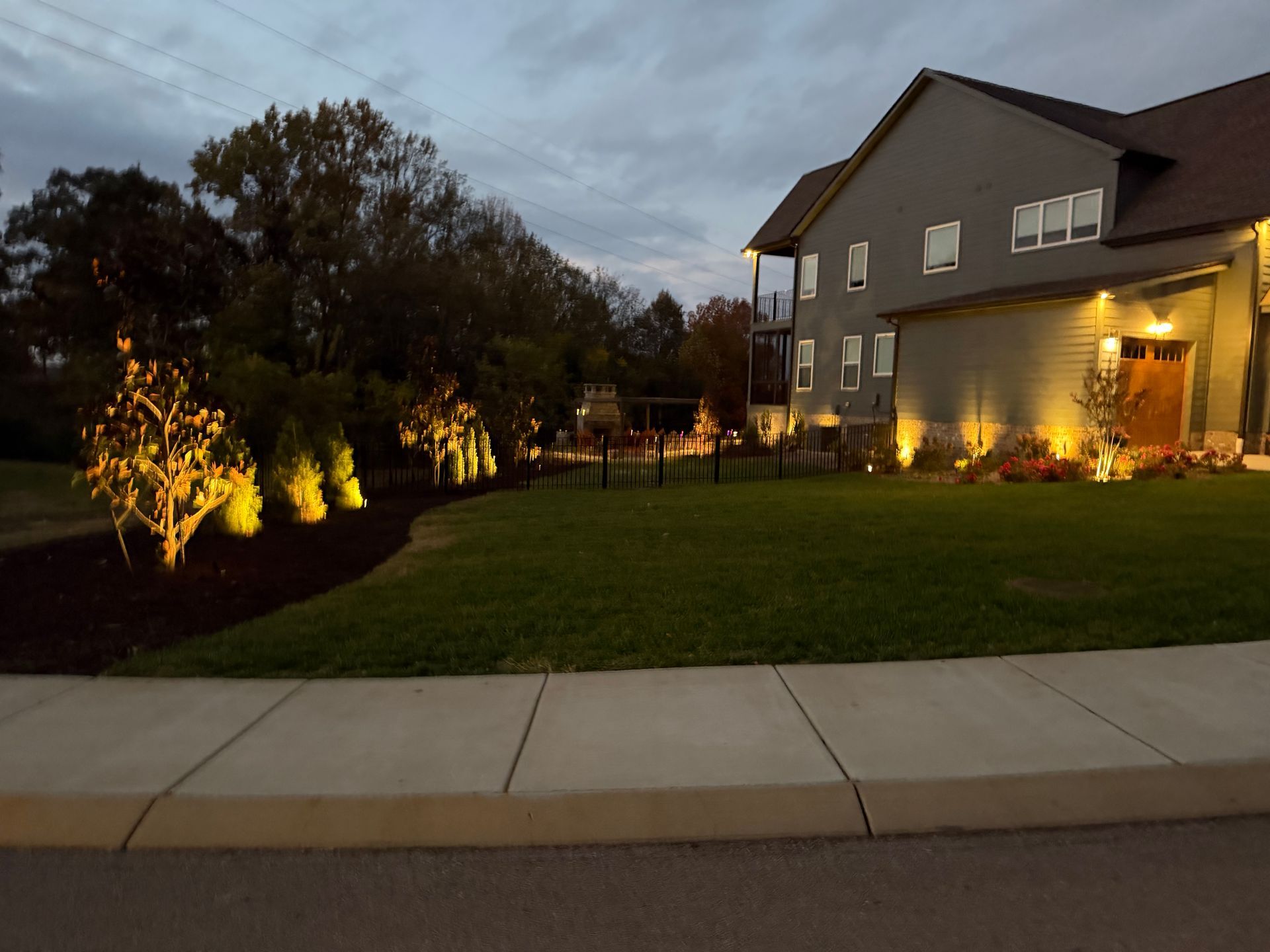 Evening view of a gray building with outdoor lighting illuminating the lawn and trees along the sidewalk.