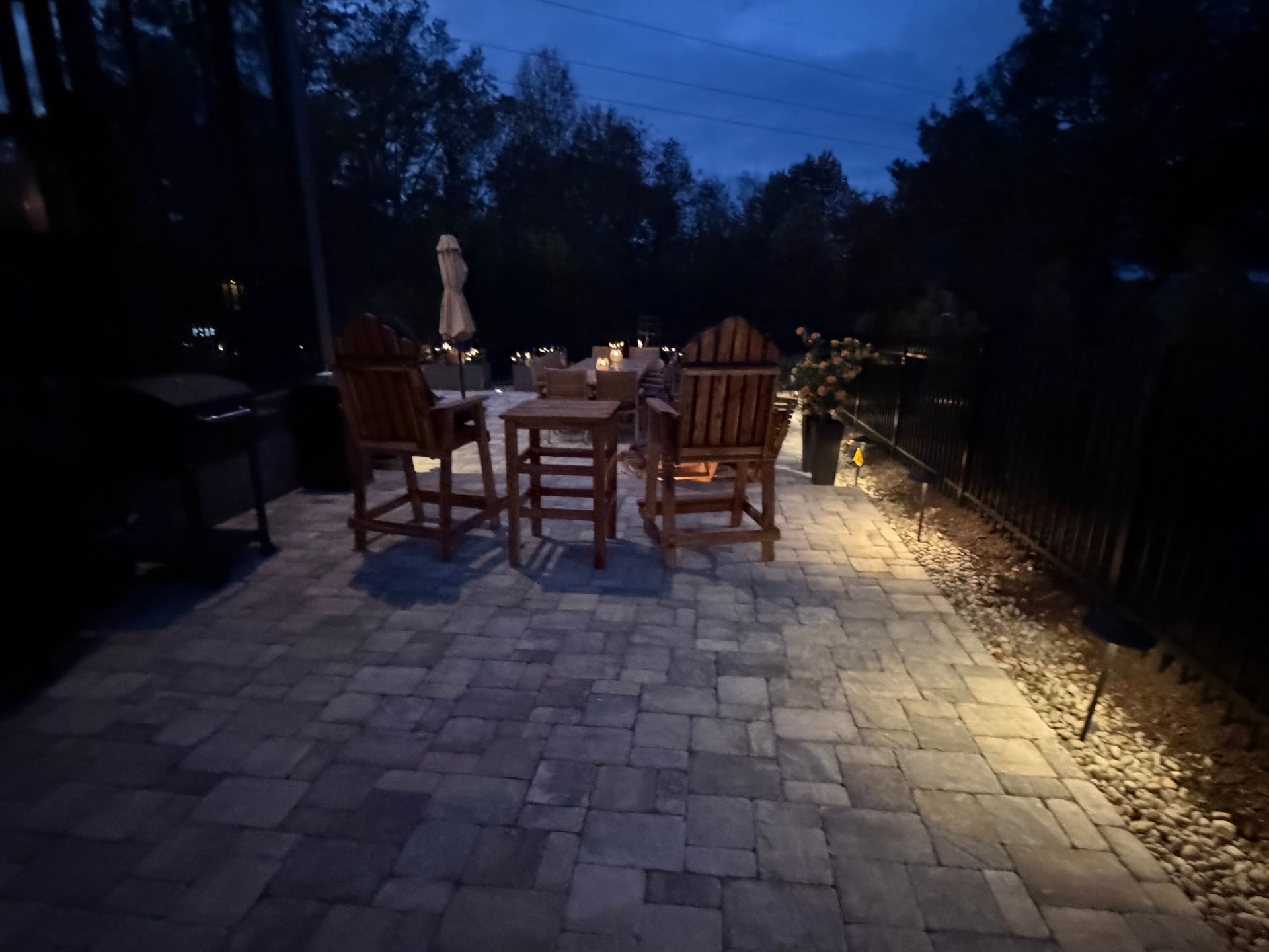 Nighttime patio scene with wooden furniture, lit by pathway lights, trees in background.