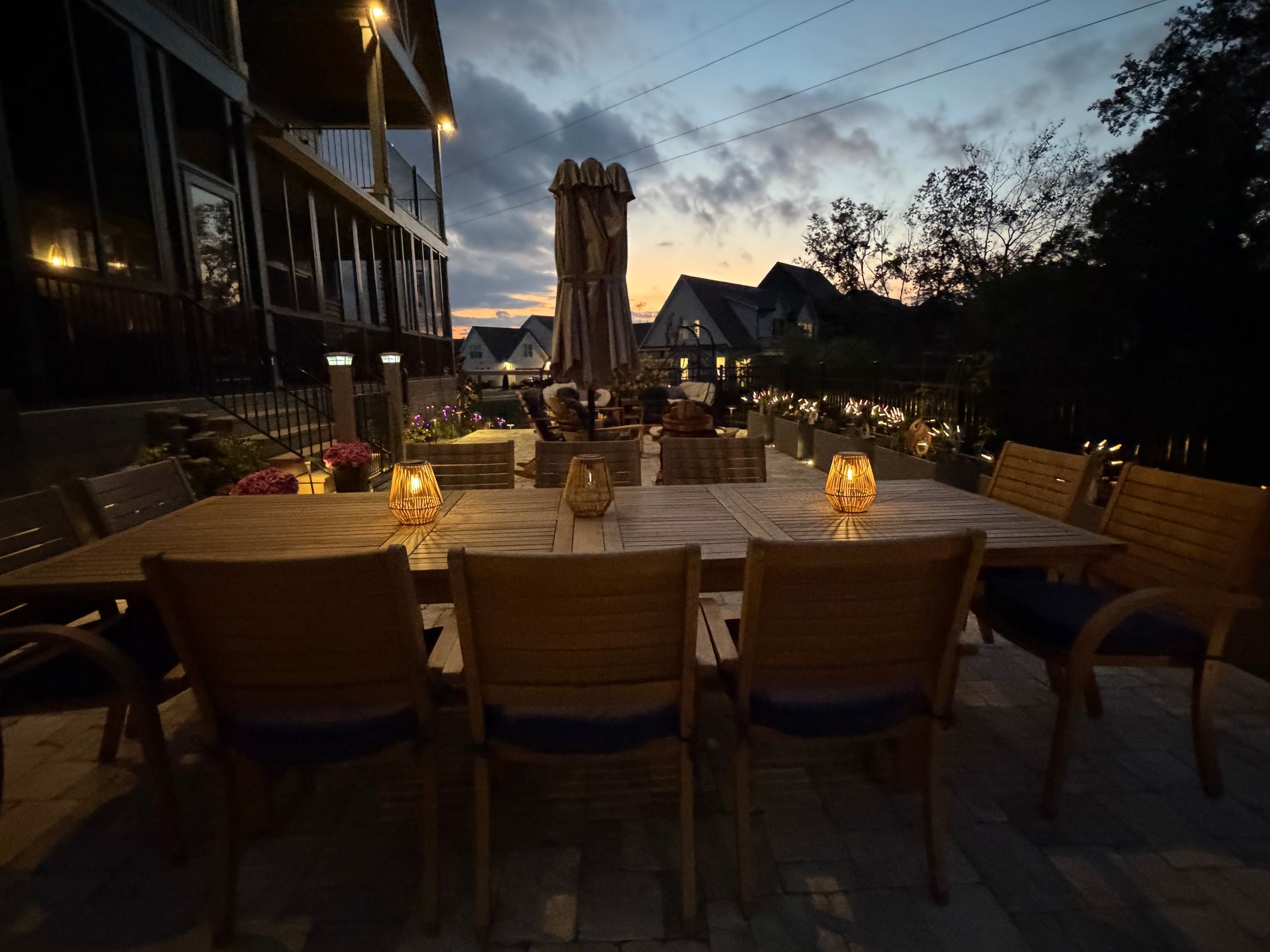 Outdoor dining table set with chairs, lit by candles at dusk, house in background.