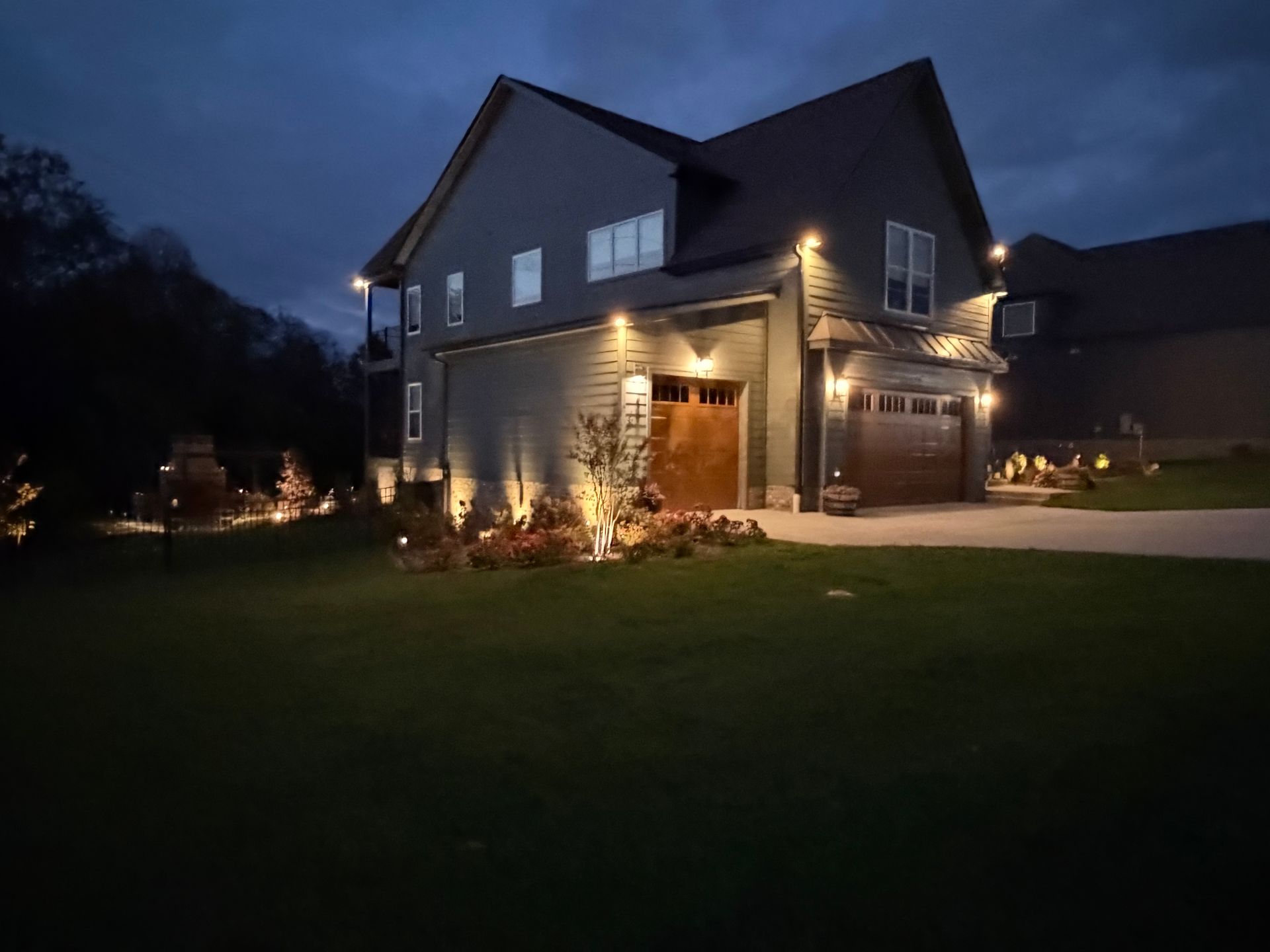 Two-story house illuminated at night. Dark blue siding, brown garage door, and landscape lighting on the lawn.