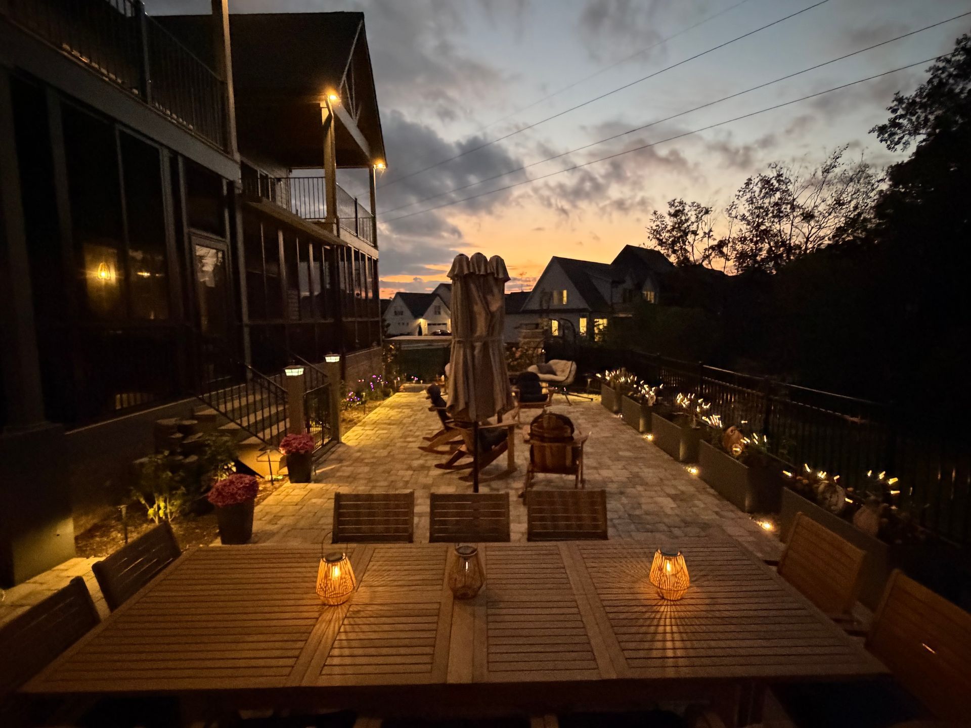 Outdoor patio with dining table, chairs, and lights at dusk.  Buildings and trees in the background.