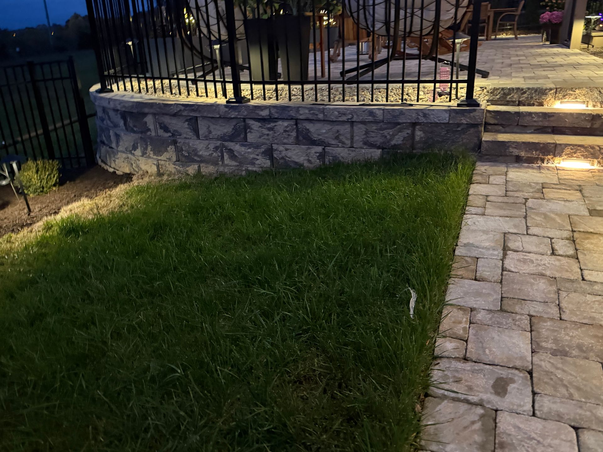Stone wall with railing, grass, and a paved walkway, illuminated at night.