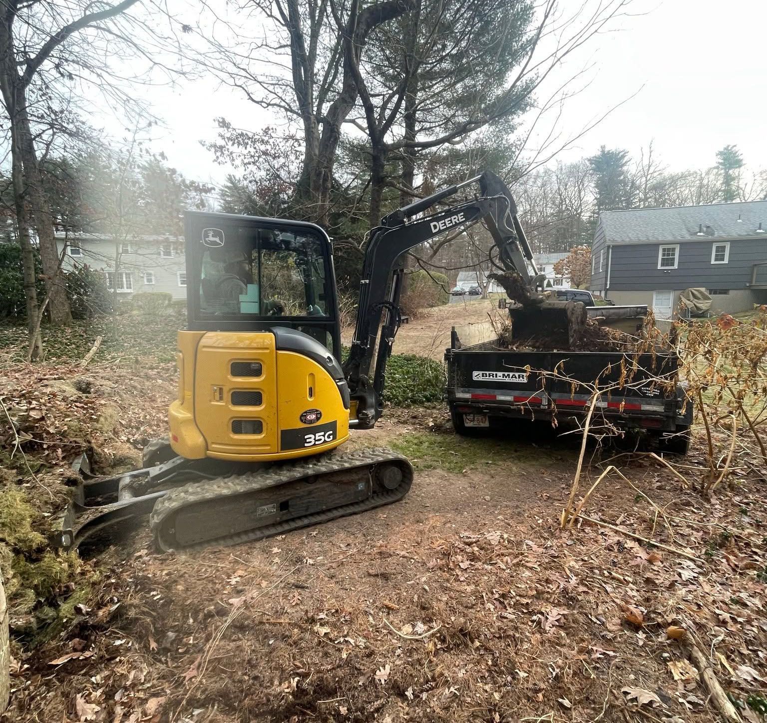 Yellow mini excavator loading debris into a black dump trailer in a leaf-covered yard under overcast skies