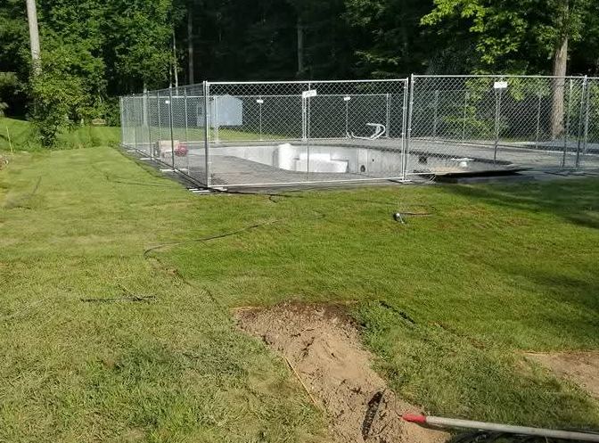 Chain-link fenced utility area beside a grassy yard, with a hole and hose in the foreground.