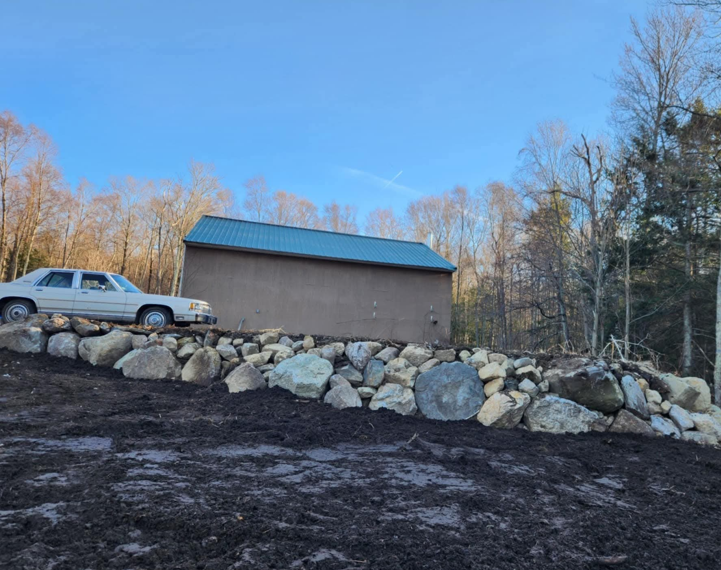 Silver pickup truck beside a tan building with blue roof, backed by a large rock retaining wall and bare trees.