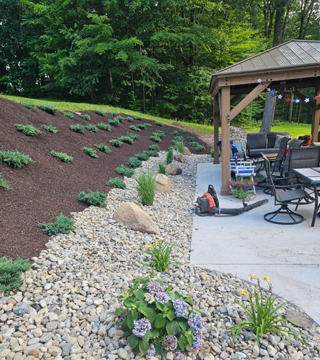 Landscaped backyard with stone path, mulch beds, young plants, and a patio gazebo with outdoor chairs