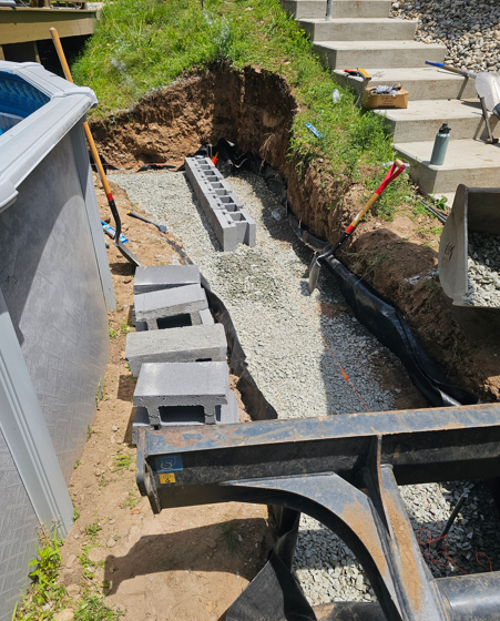 Excavated trench with gravel base, cinder blocks, and black liner beside a yard retaining wall