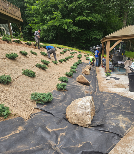 Workers installing plants and black landscape fabric on a sloped garden hillside near a gazebo.