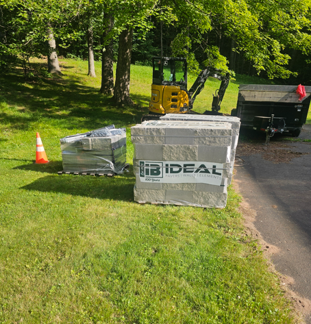 Construction materials and equipment on a grassy driveway beside trees, with an IDEAL box and black dumpster.