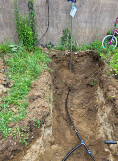 Deep trench dug in a backyard, with exposed soil, a black hose, and a shovel near a fence.