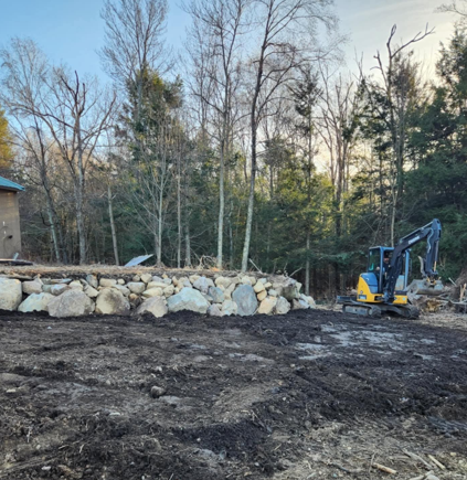 Cleared dirt lot with stacked rocks, bare trees, and a small excavator at the right edge