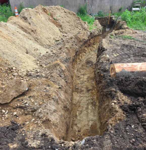 A long narrow trench dug in a dirt yard beside a soil mound and exposed pipe.